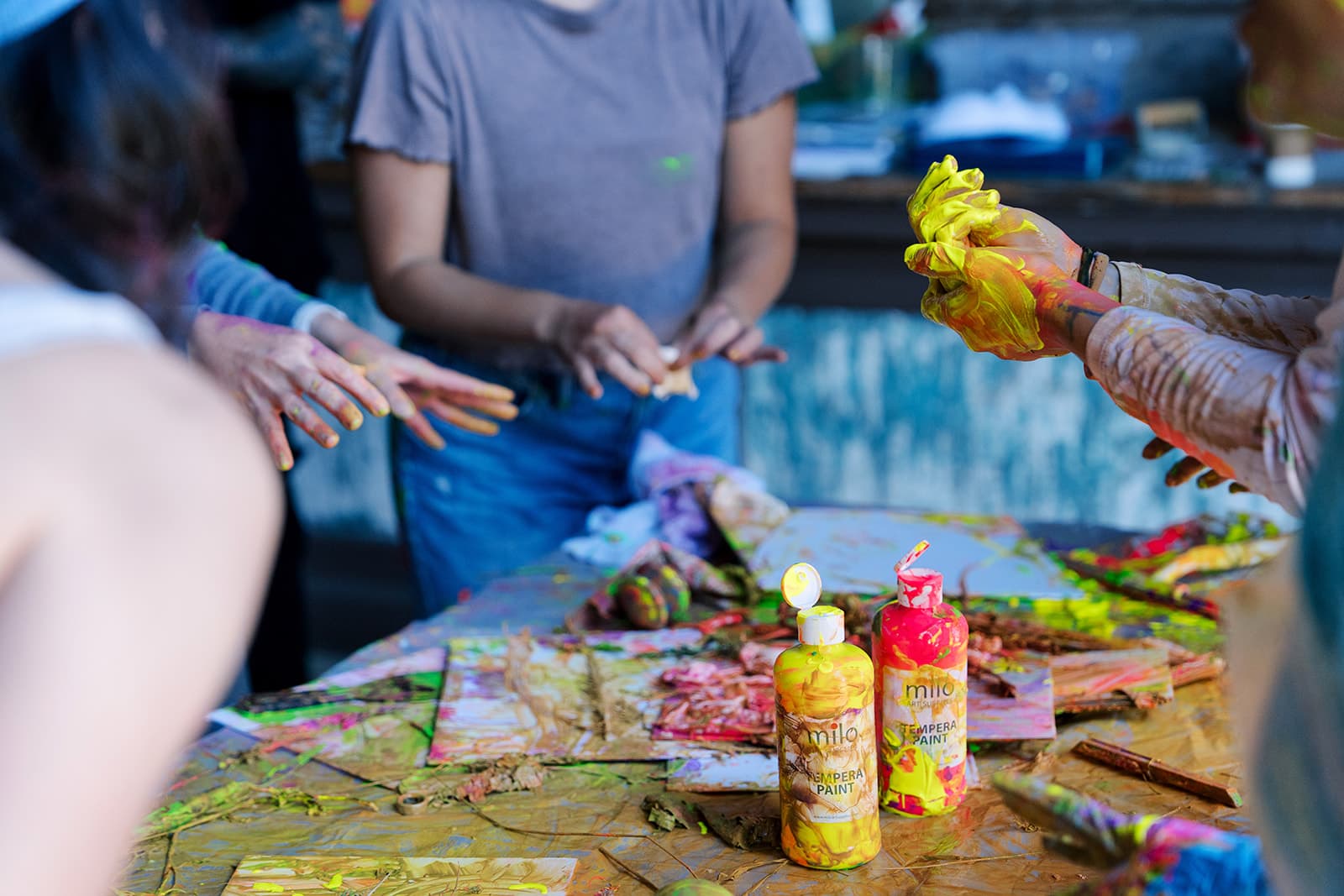 Paint-covered hands reaching across a table of tempera bottles