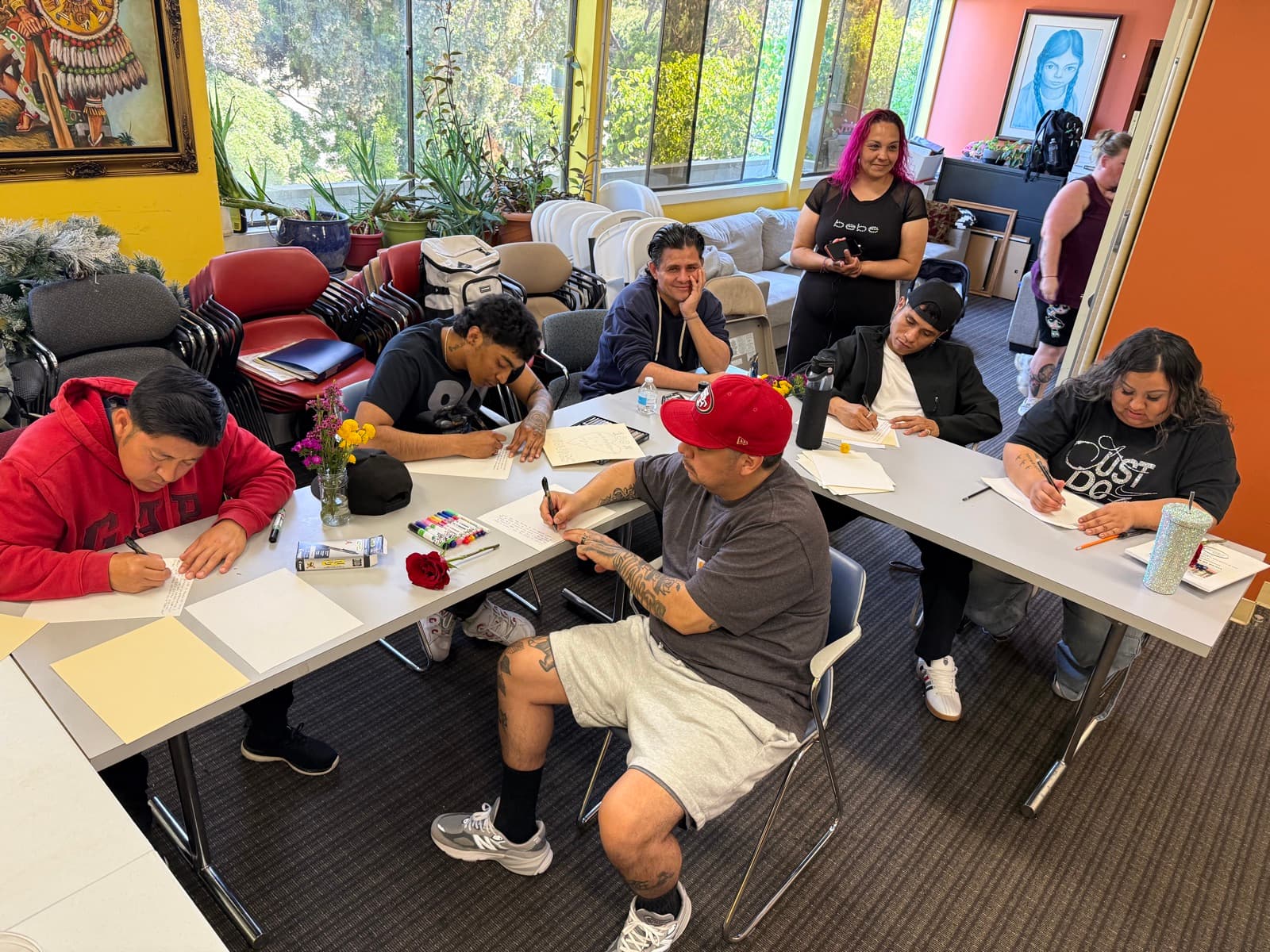 Residents seated around tables writing and drawing, with flowers in jars and an Aztec warrior painting on the yellow wall behind them