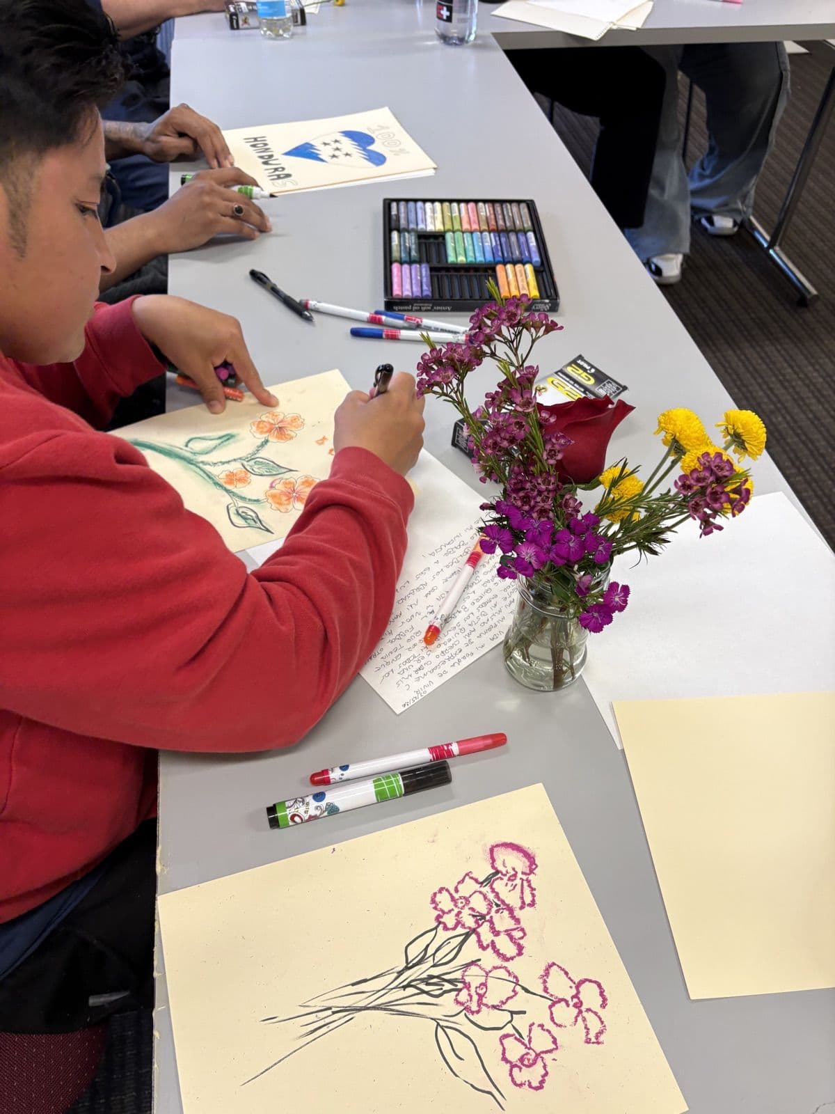 A resident drawing orange flowers in oil pastel beside a jar of fresh flowers and a tray of pastels