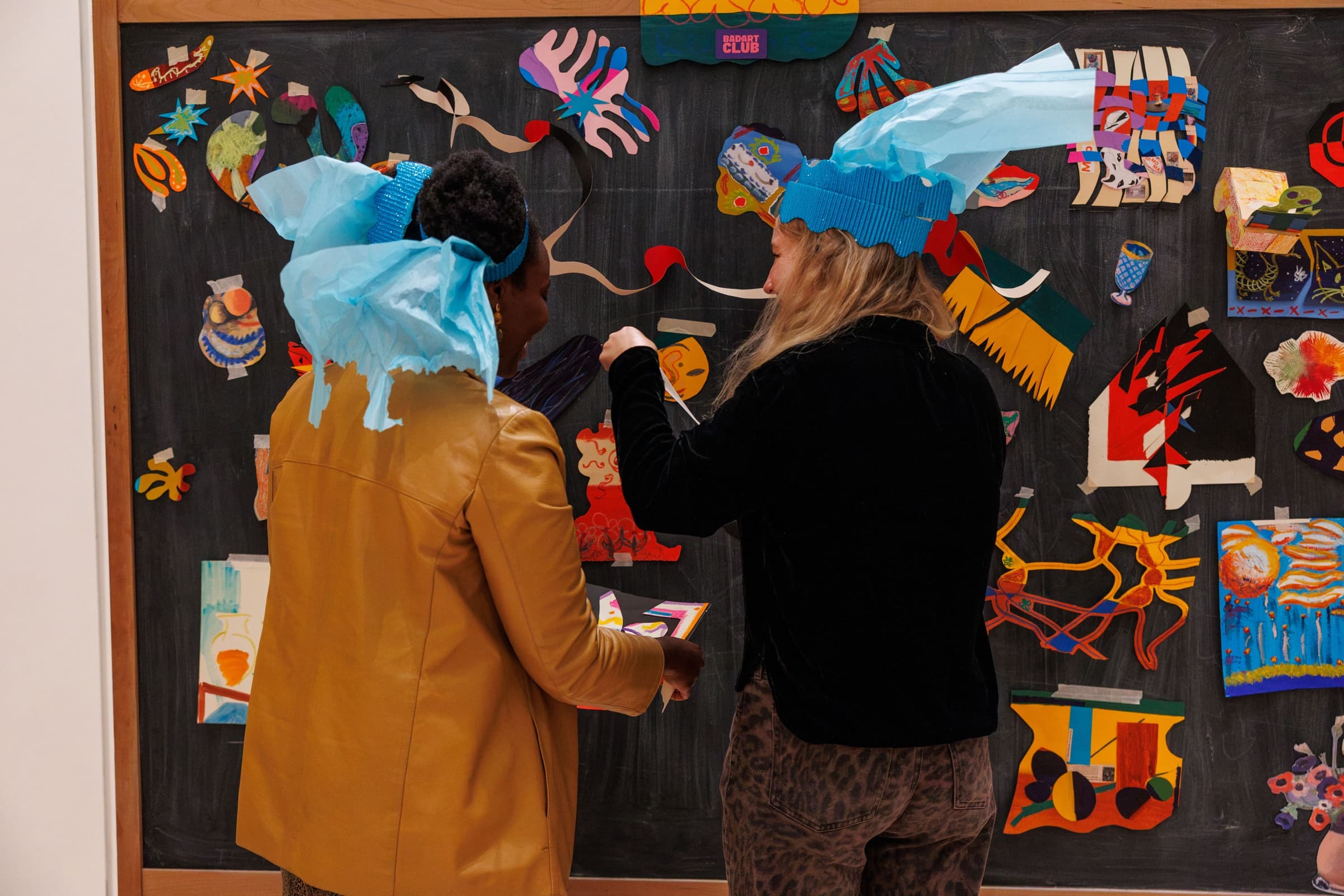 Two participants in handmade blue paper hats adding pieces to a wall-sized collaborative collage