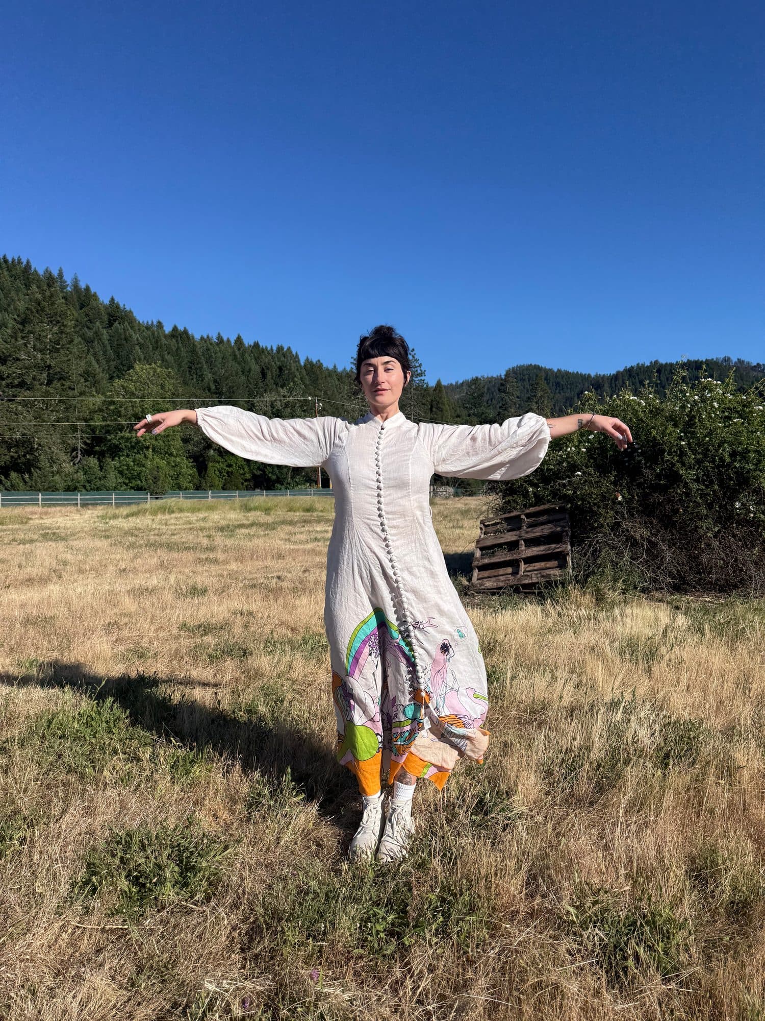 Julianne Costa standing arms wide in a sunlit meadow beneath the mountains.