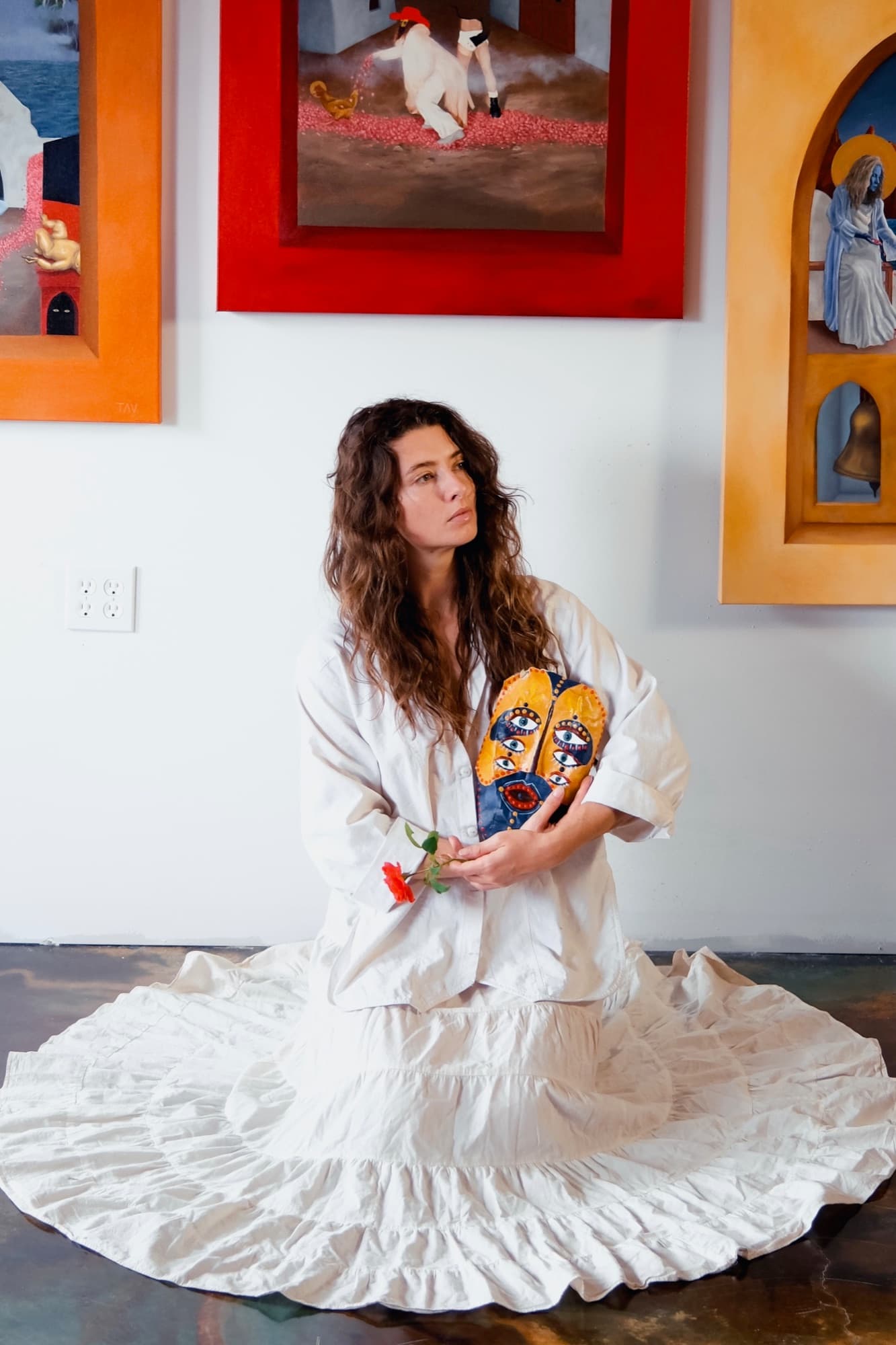 Taylor Ana Valdez in a white dress, holding a painted mask in her gallery.