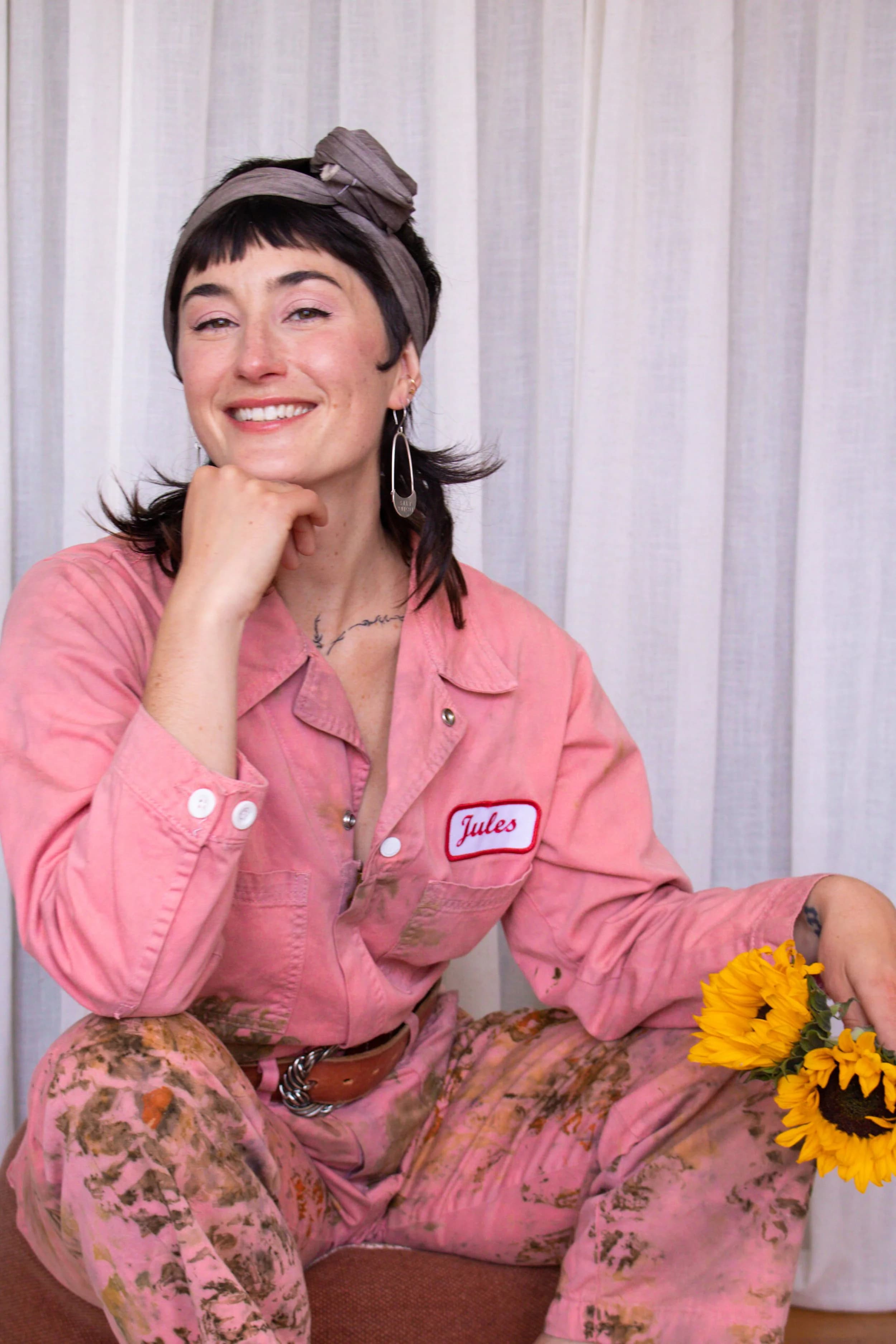 Jules in pink jumpsuit with headscarf and earrings smiling, holding sunflowers.