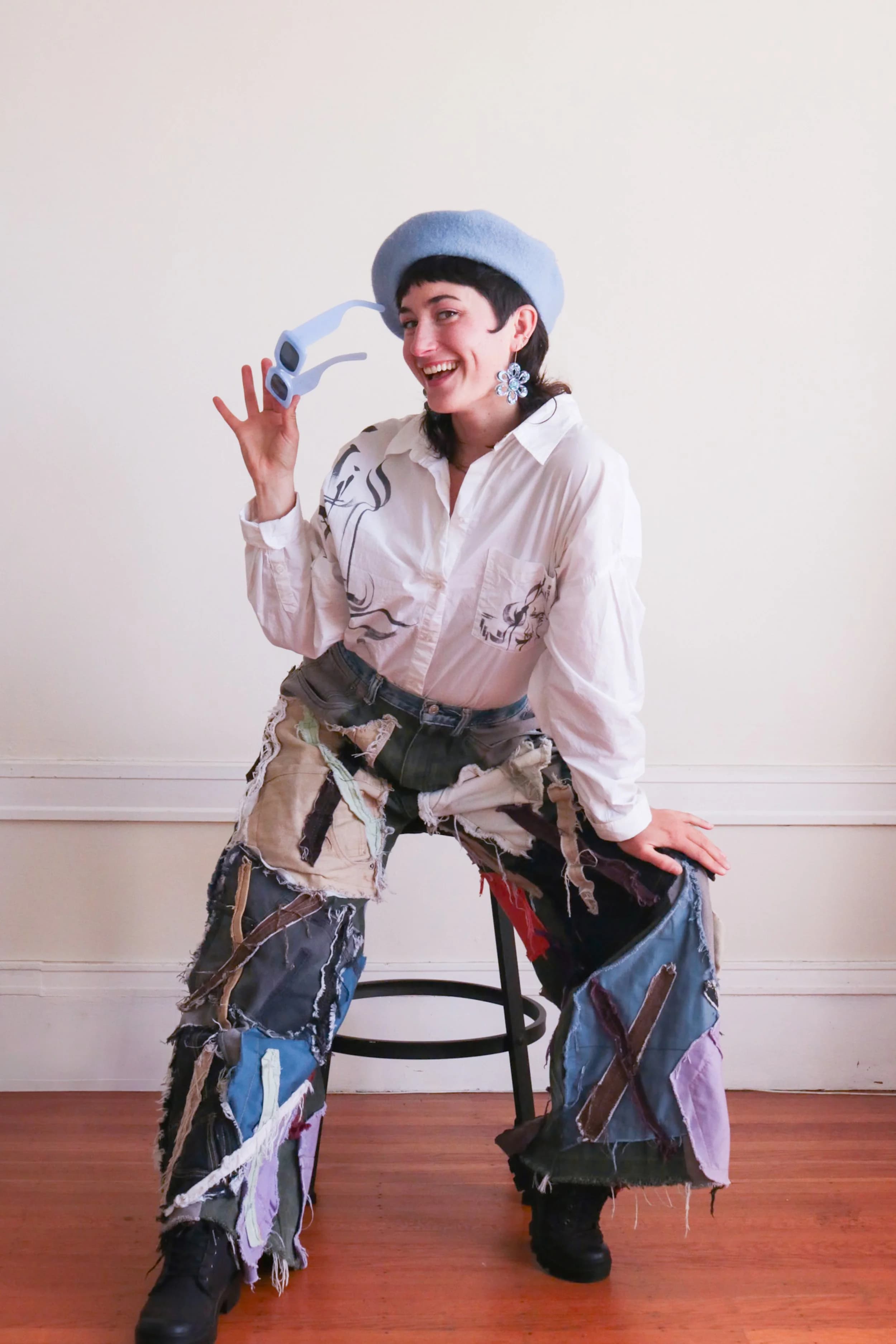 Jules wearing a blue beret, white shirt, patched wide-leg pants, holding blue sunglasses, sitting on a stool inside a room with wooden floor.