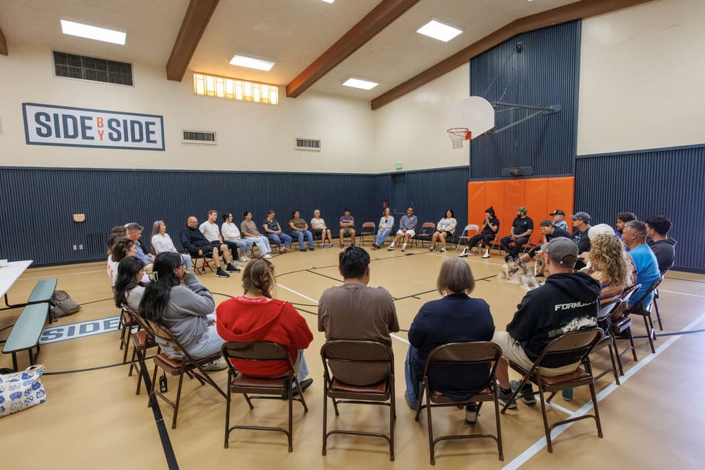 38 Hunt School staff seated in a large circle in the gymnasium beneath the Side By Side logo