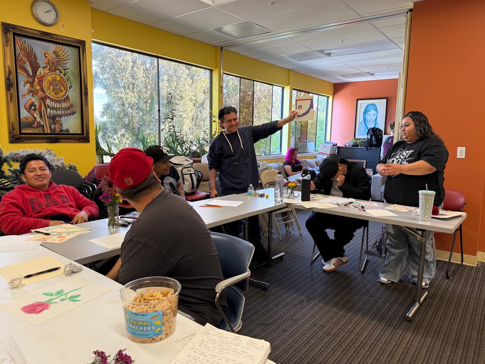 A resident smiling and holding up his artwork to the group during show and tell, in a sunny room with yellow walls