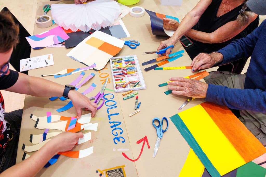 Many hands reaching across a table covered in colorful cut paper, scissors and pastels, working on a collaborative collage