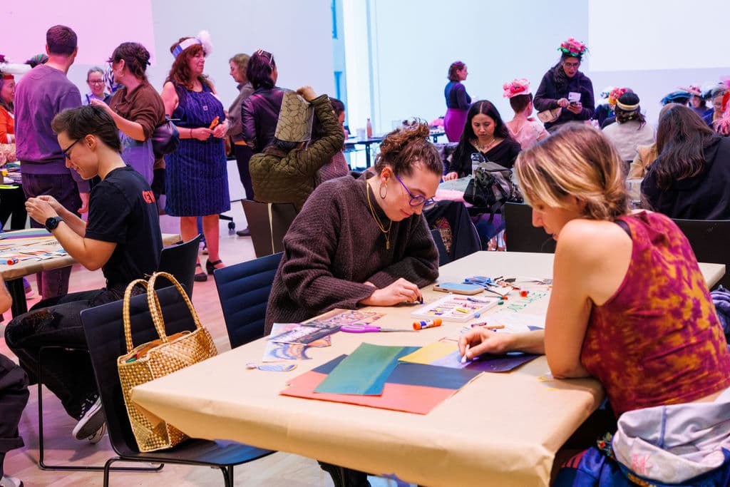 A packed room at SFMOMA with people at art-making tables, handmade hats on their heads, and a large collaborative collage covering the back wall