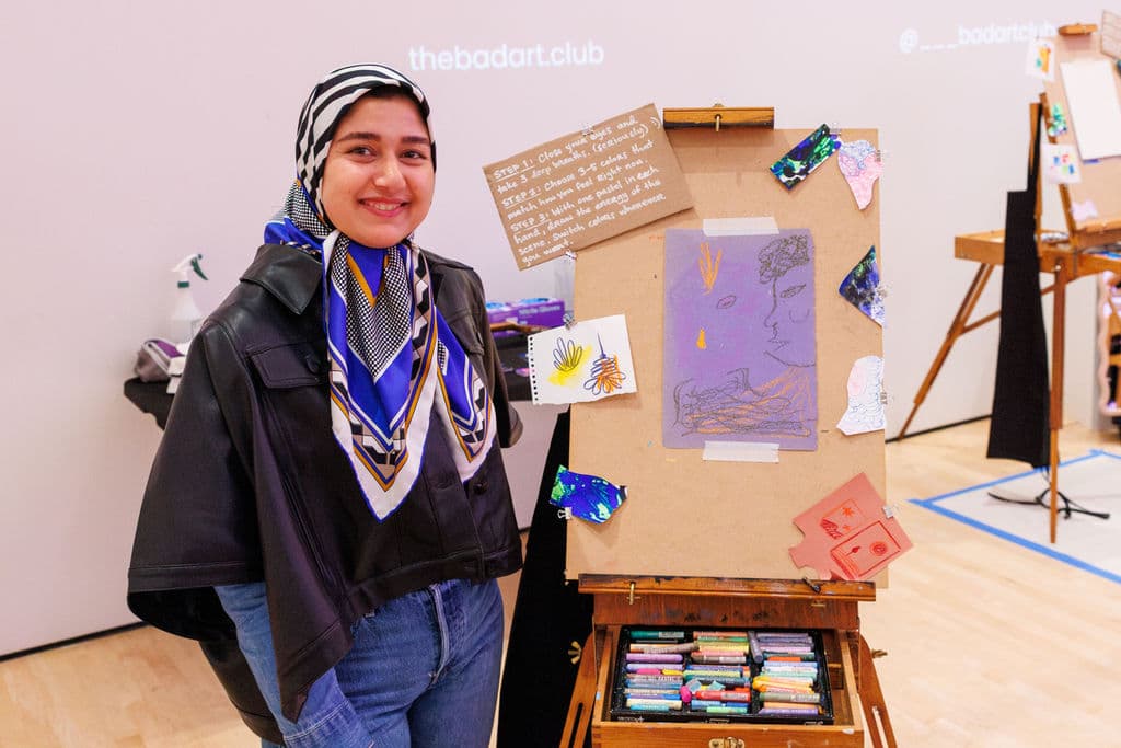 A participant smiling proudly beside her finished painting on an easel, the Bad Art Club projection visible behind her