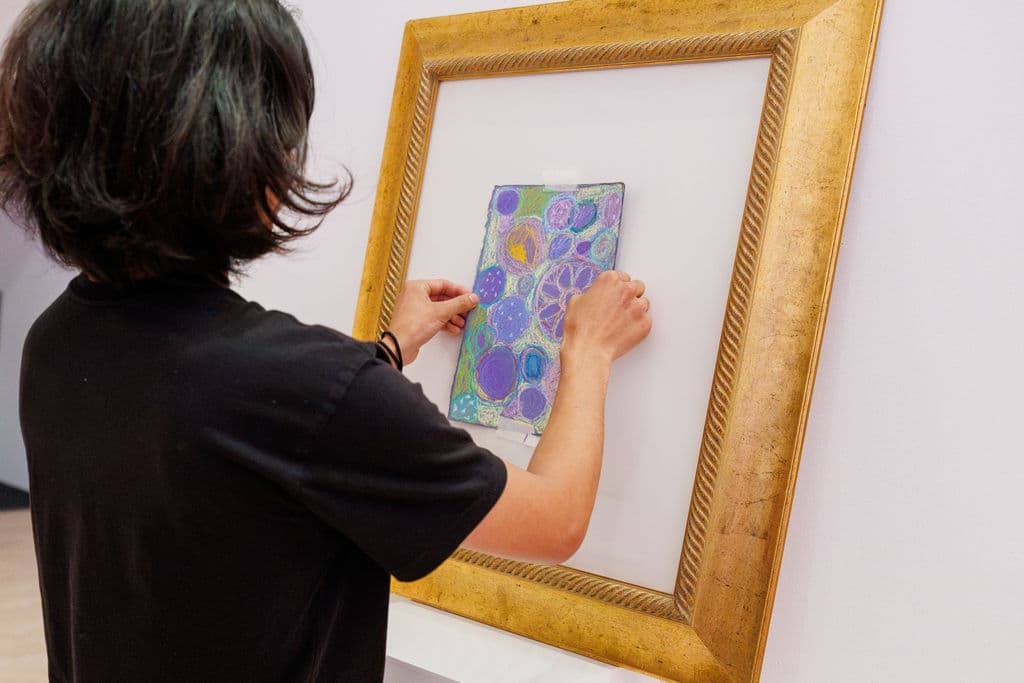 A participant carefully placing their pastel painting into a large gilded museum frame mounted on the gallery wall