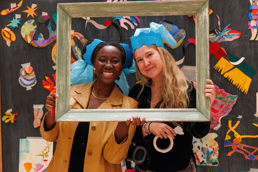 Two participants with handmade blue paper hats holding an empty ornate picture frame around their faces and laughing