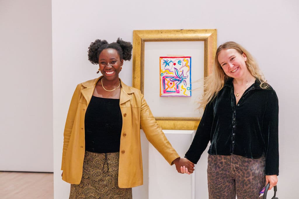 Two women holding hands and smiling in front of a gold-framed artwork displayed on a pedestal in the SFMOMA gallery