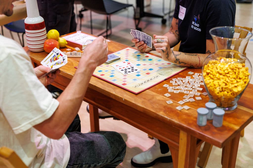 People playing cards and Scrabble together at a table, a Bad Art Club shirt visible in the background
