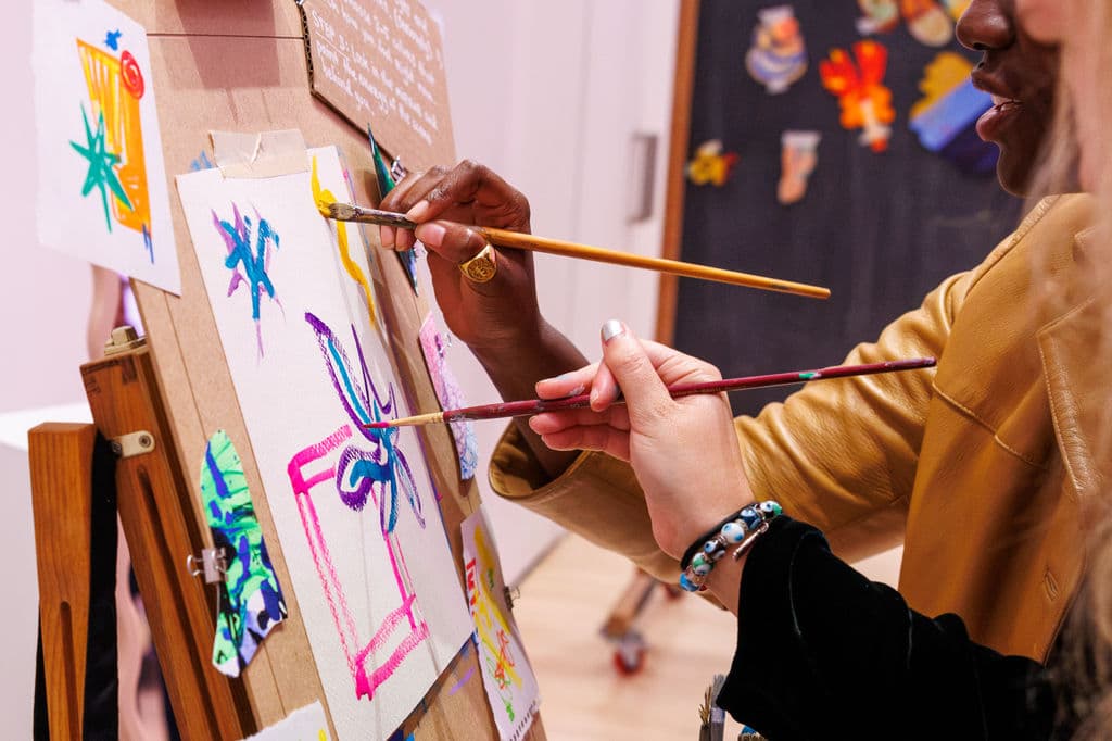 Two hands, one with a gold ring and one with a bracelet, both holding brushes and painting the same canvas at an easel