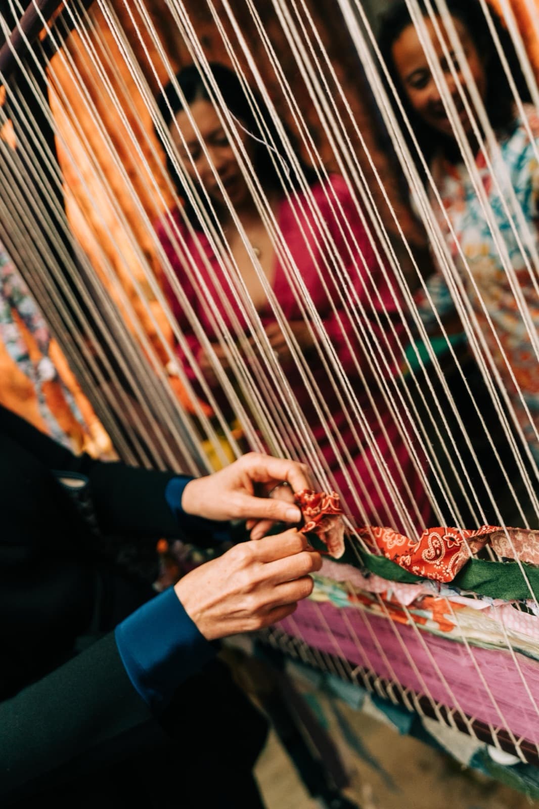Close-up of hands knotting a red paisley strip into the loom, two guests smiling through the warp threads behind