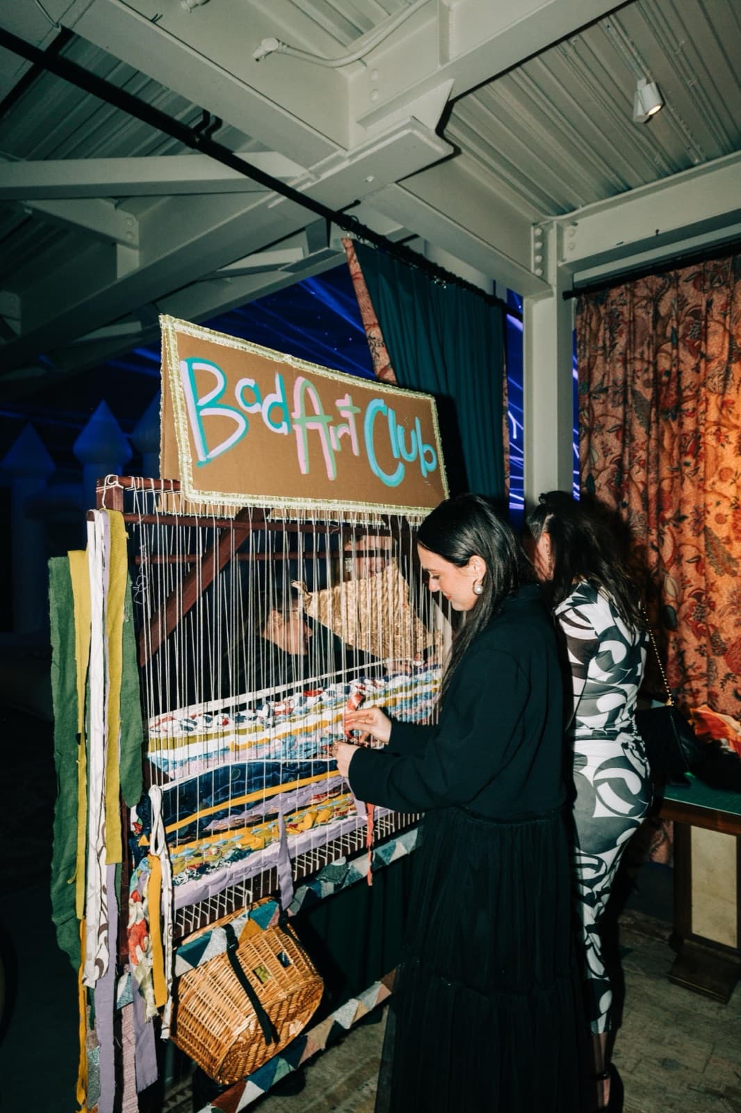 A guest weaving fabric into the Bad Art Club loom beneath a hand-painted cardboard Bad Art Club sign, a basket of fabric scraps hanging below