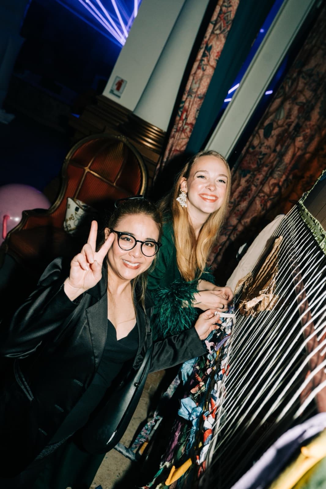 Two guests smiling at the loom, one flashing a peace sign, beneath the ornate curtains and blue lights of the venue