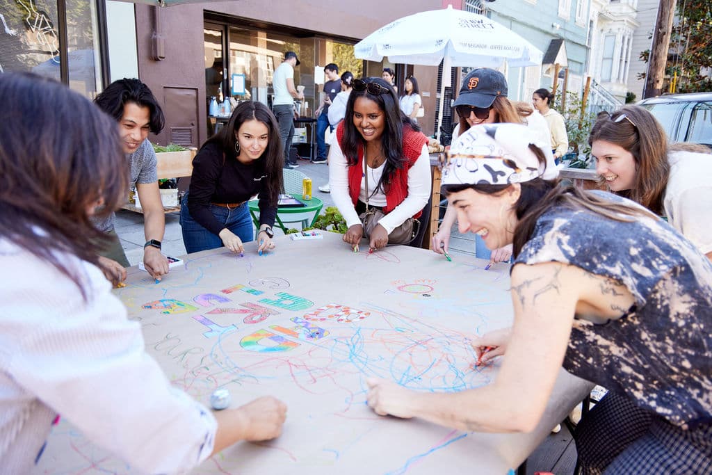 A group of people gathered around a large table making collaborative art outdoors