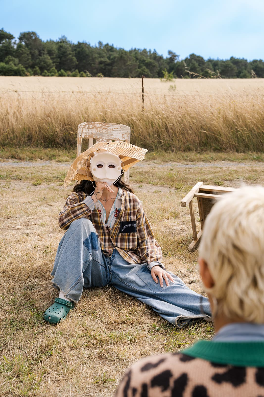 A person wearing a white mask and straw hat sits in a golden field during a Bad Art Club workshop