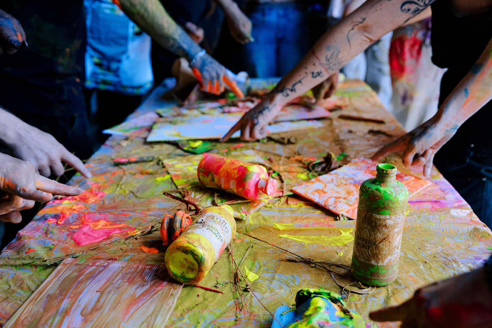 A chaotic table covered in neon paint, bottles, and hands reaching in